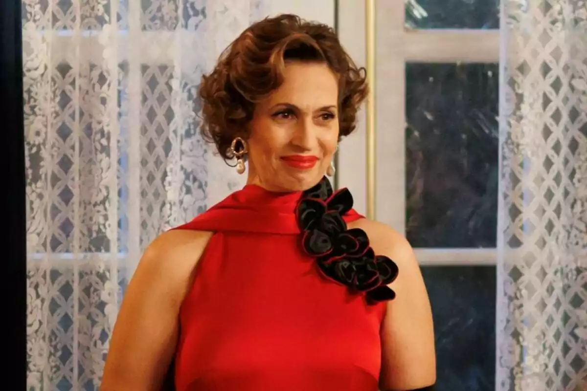 Lola Marceli con un vestido rojo elegante y detalles florales negros sonríe frente a una ventana con cortinas de encaje en La Moderna. Lola Marceli con un vestido rojo elegante y detalles florales negros sonríe frente a una ventana con cortinas de encaje en La Moderna.