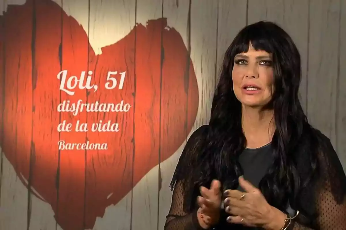 Loli Sánchez de cabell llarg i fosc amb vestit negre parlant davant d’una paret de fusta clara amb un gran cor vermell i text que diu Loli 51 gaudint de la vida Barcelona a First Dates