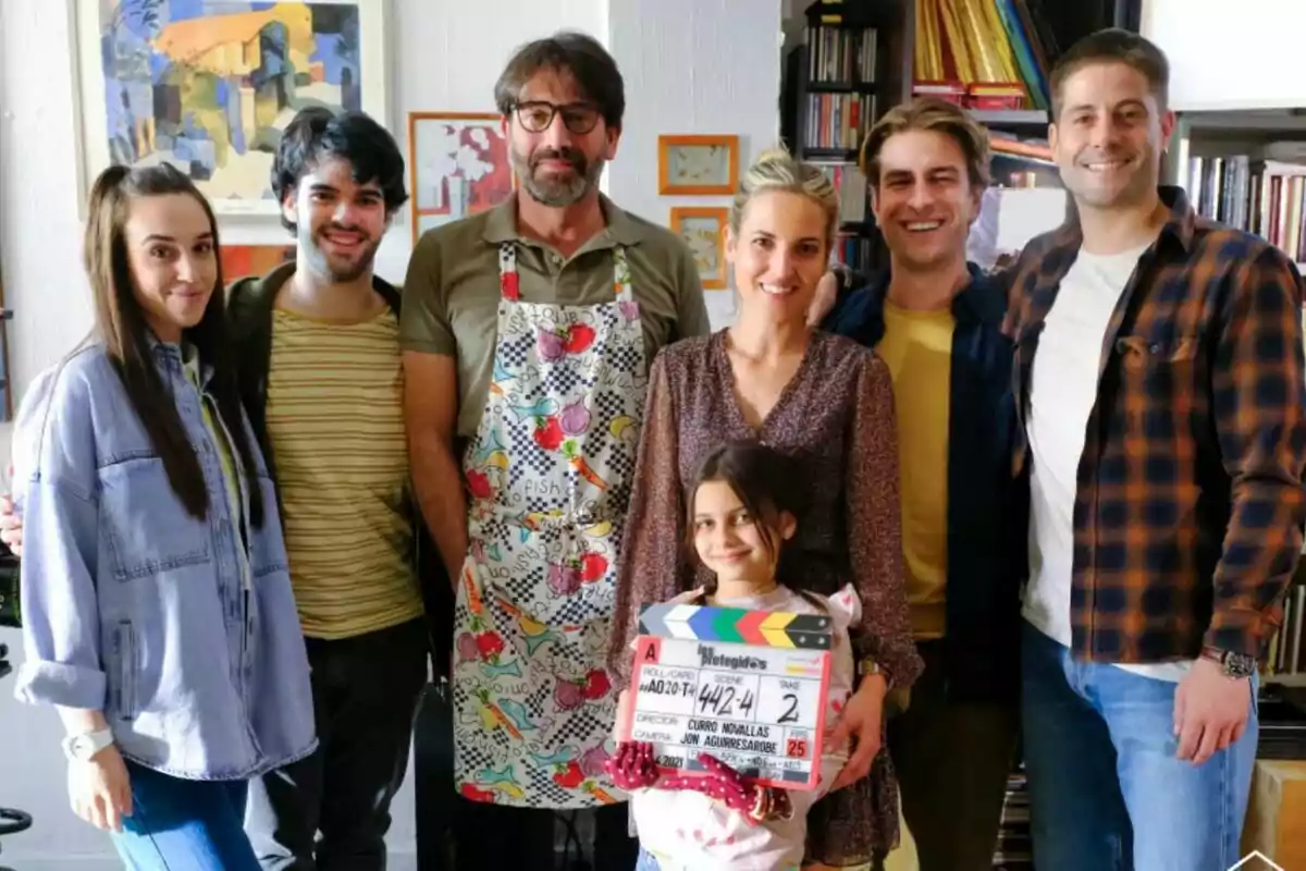Un grupo de personas como Ana Fernández, Luis Fernández, Mario Marzo o Antonio Garrido sonríe mientras Cosette Silguero sostiene una claqueta de cine en un ambiente acogedor con libros y cuadros en el fondo en Los Protegidos.