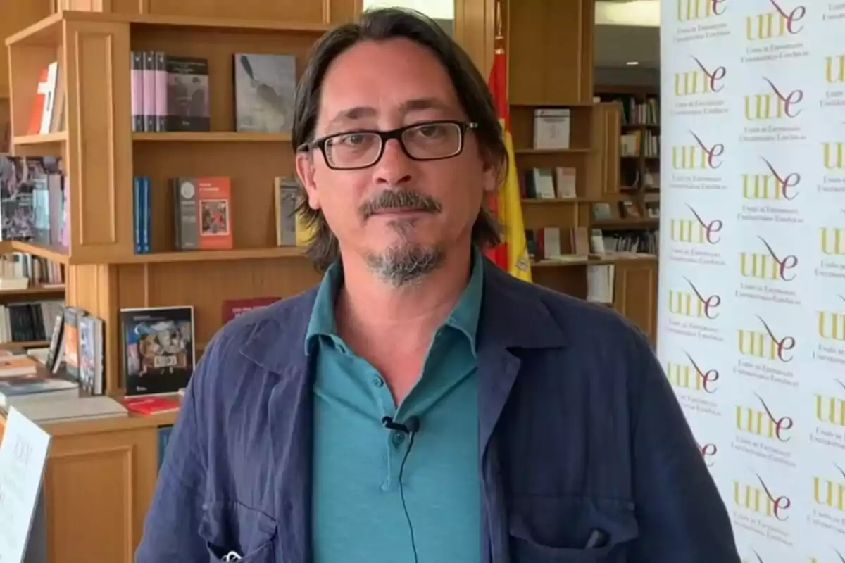 Luis Felipe Torrente amb ulleres i barba està dret en una biblioteca, amb prestatgeries plenes de llibres darrere seu i un cartell de la Unió d'Editorials Universitàries Espanyoles (UNE) a la dreta. Luis Felipe Torrente amb ulleres i barba està dret en una biblioteca, amb prestatgeries plenes de llibres darrere seu i un cartell de la Unió d'Editorials Universitàries Espanyoles (UNE) a la dreta.