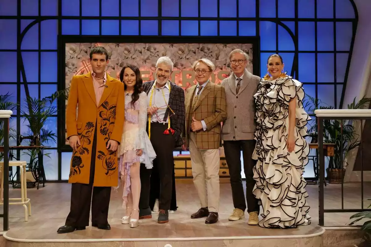 El jurat de 'Maestros de la Costura Celebrity' posant al costat dels dissenyadors Victorio & Luchino i al costat de la presentadora Raquel Sánchez Silva, cadascun amb vestits elegants i acolorits, amb un fons de panells blaus i plantes decoratives.