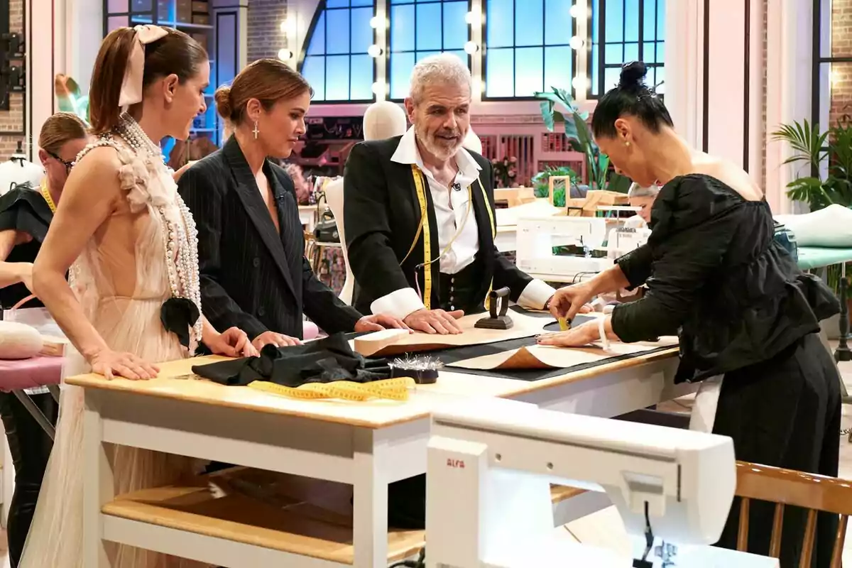 Lorenzo Caprile, Adriana Ugarte i Raquel Sánchez Silva estan reunits al voltant d'una taula de treball en un taller de costura, observant i col·laborant en un projecte de confecció de Rosa López a Maestros de la Costura.