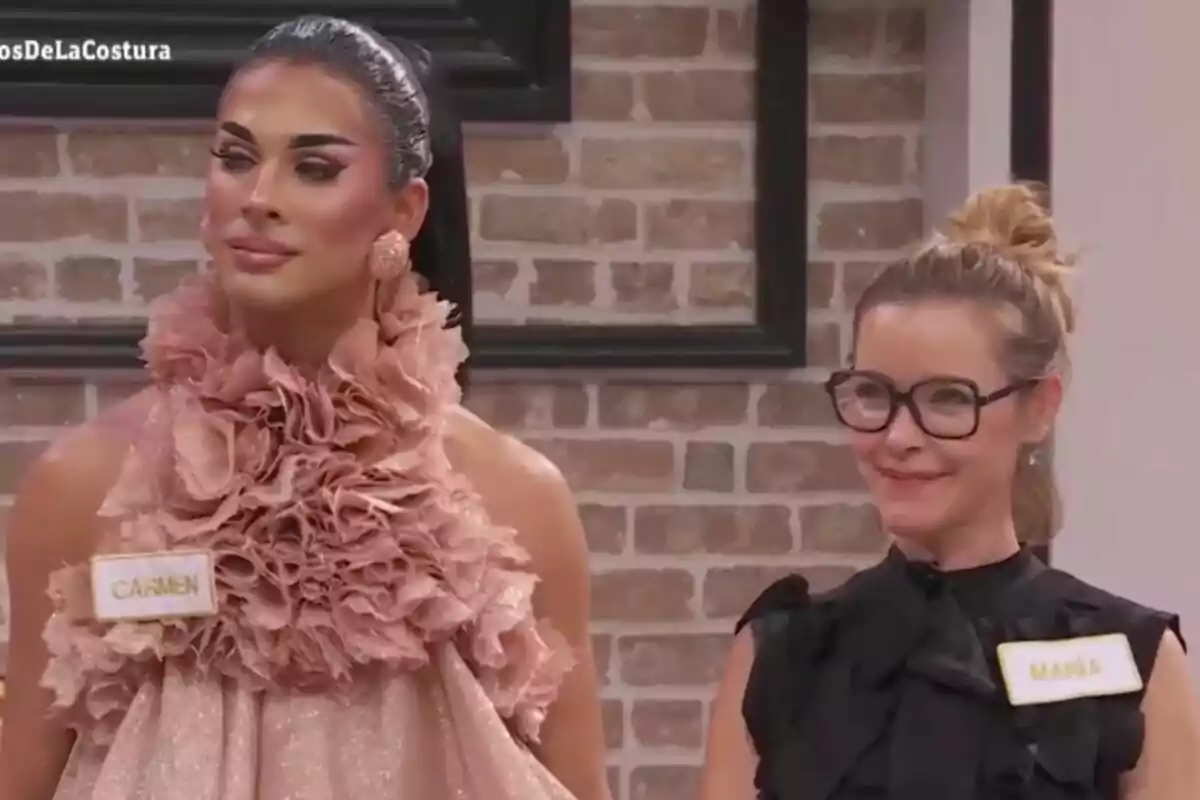 Carmen Farala y María Esteve posan juntas en un entorno interior con paredes de ladrillo, una lleva un vestido con volantes y la otra viste de negro y usa gafas en 'Maestros de la Costura'.