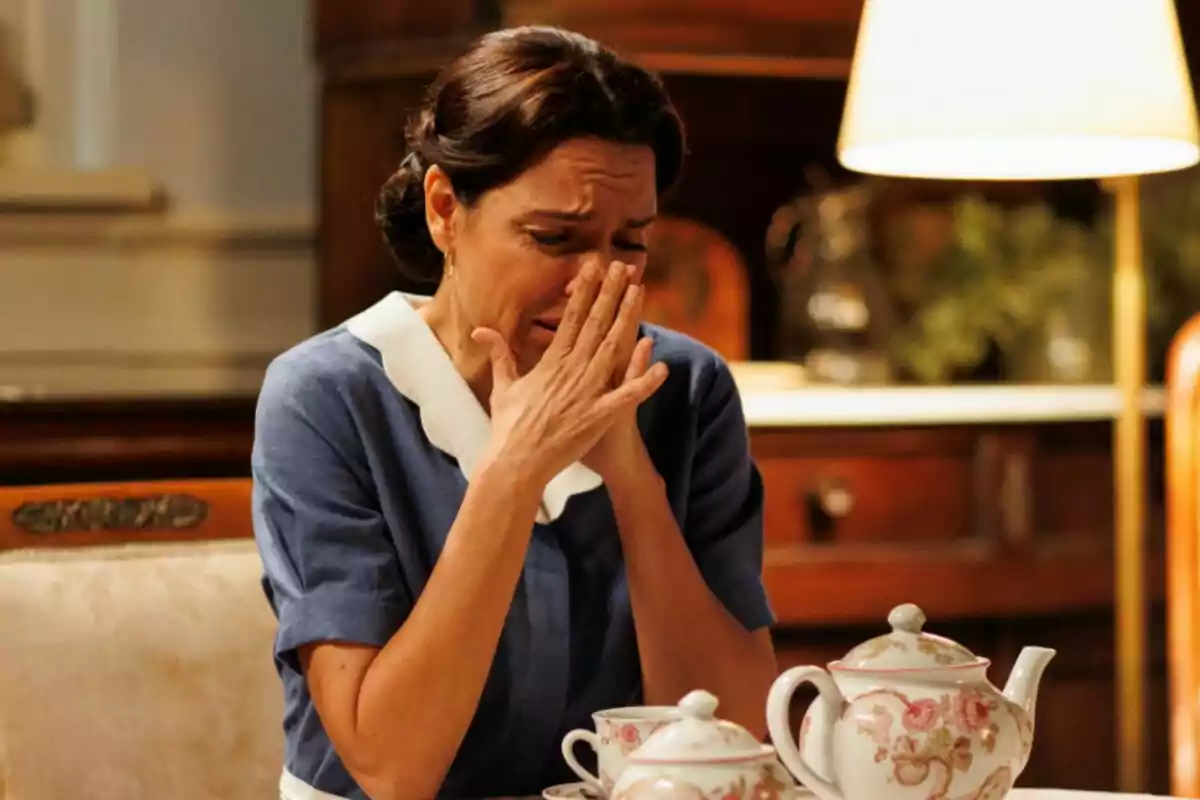 Magdalena Tejado con uniforme azul llorando con las manos en el rostro sentada frente a una mesa con un juego de té en La Moderna. Magdalena Tejado con uniforme azul llorando con las manos en el rostro sentada frente a una mesa con un juego de té en La Moderna.