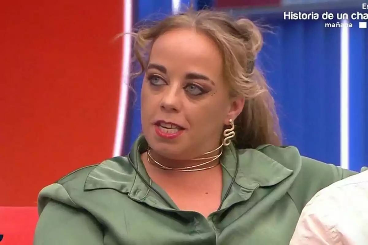 Maite amb brusa verda i accessoris daurats està asseguda en un sofà vermell amb un fons blau a Gran Hermano. Maite amb brusa verda i accessoris daurats està asseguda en un sofà vermell amb un fons blau a Gran Hermano.