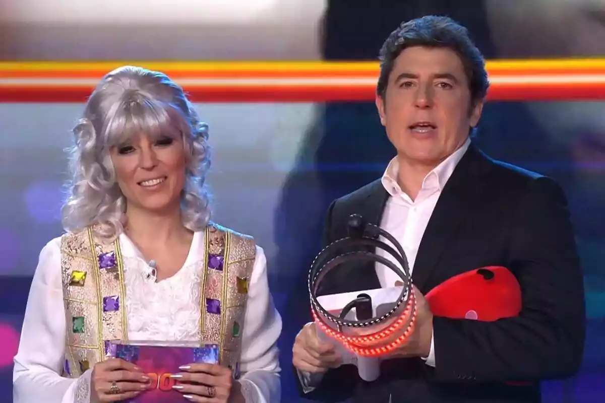 Dos personas en un escenario, Gisela con peluca plateada y chaleco decorado sostiene un sobre, mientras Manel Fuentes con traje oscuro sostiene un objeto con luces rojas en Tu Cara Me Suena.