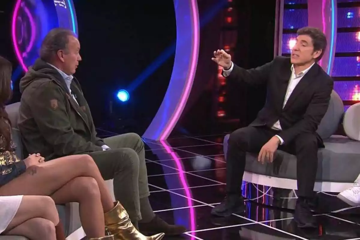 Bertín Osborne junto a Manel Fuentes está sentado en Tu Cara Me Suena iluminado con luces de colores, mientras un hombre de traje gesticula durante una conversación. Bertín Osborne junto a Manel Fuentes está sentado en Tu Cara Me Suena iluminado con luces de colores, mientras un hombre de traje gesticula durante una conversación.