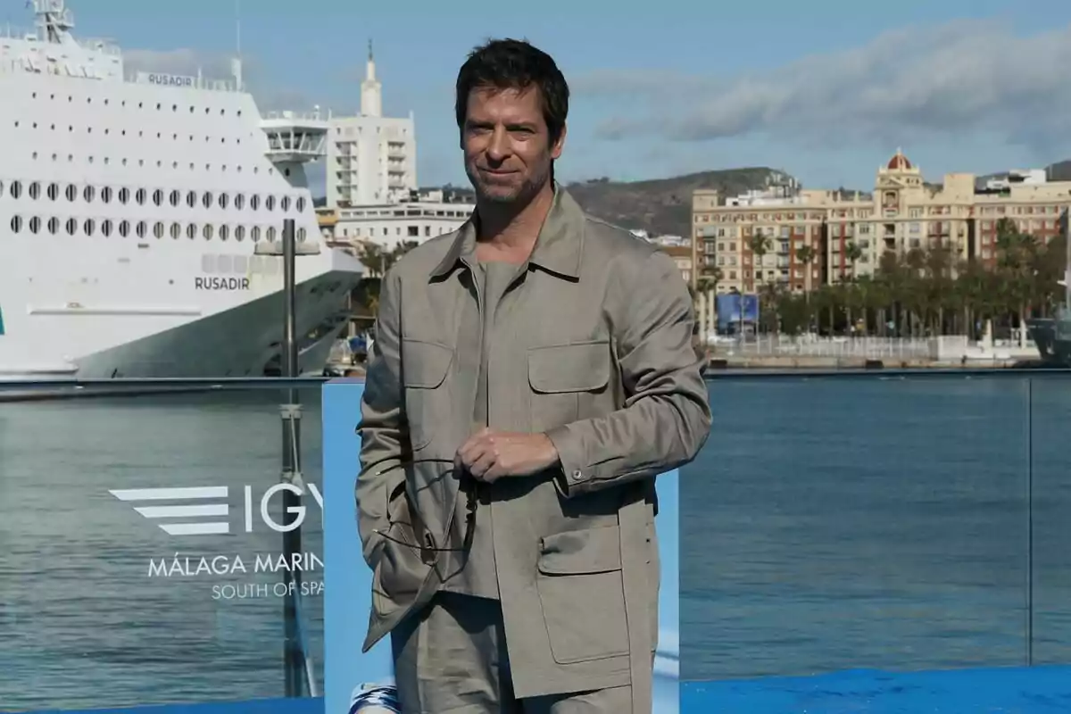 Manu Baqueiro amb vestit clar posant davant d'un port amb un gran vaixell i edificis al fons en la presentació de Perdiendo el Juicio.