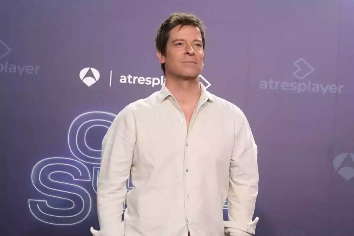 Manu Baqueiro amb camisa blanca posant davant d'un fons amb logotips de Tu Cara Me Suena i Atresplayer.
