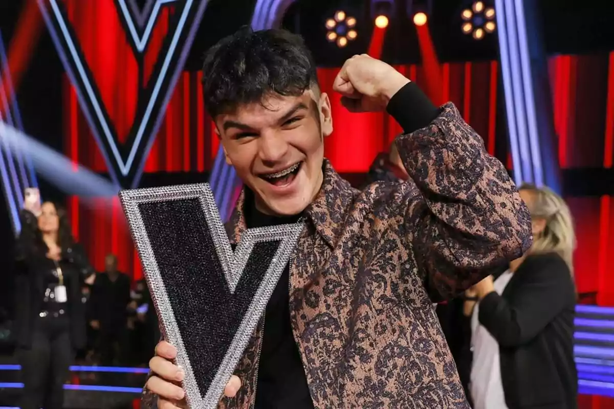 Manuel Ayra sonriente levanta un trofeo de La Voz en forma de V en un escenario iluminado con luces rojas y azules. Manuel Ayra sonriente levanta un trofeo de La Voz en forma de V en un escenario iluminado con luces rojas y azules.