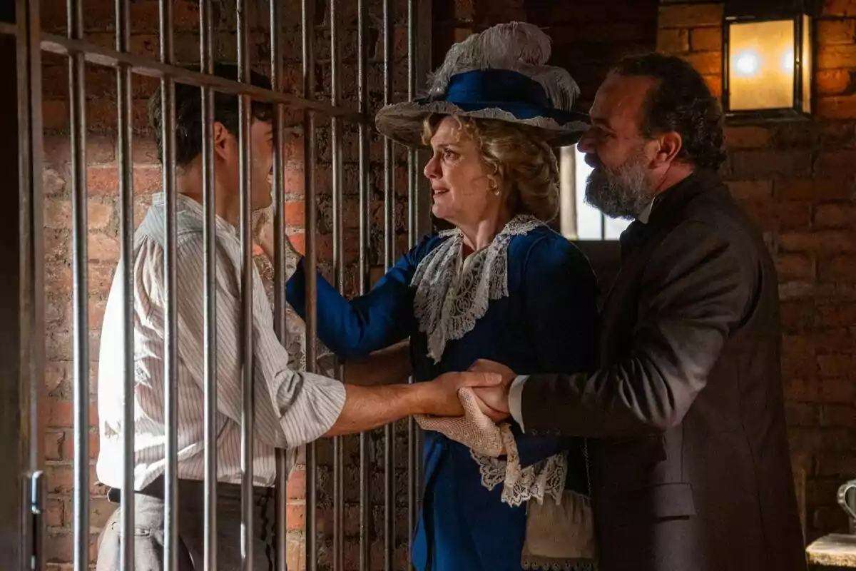 Manuel Regueiro y Eva Martín vestidos de época sostienen las manos de Arturo García Sancho tras las rejas en una celda iluminada por una lámpara en La Promesa. Manuel Regueiro y Eva Martín vestidos de época sostienen las manos de Arturo García Sancho tras las rejas en una celda iluminada por una lámpara en La Promesa.