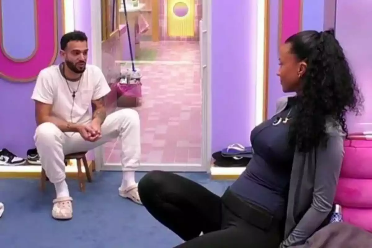 Manuel Cortés i Jeimy Báez conversant en una habitació amb parets de color porpra i rosa, una està asseguda en una cadira i l'altra a terra a GH DÚO.