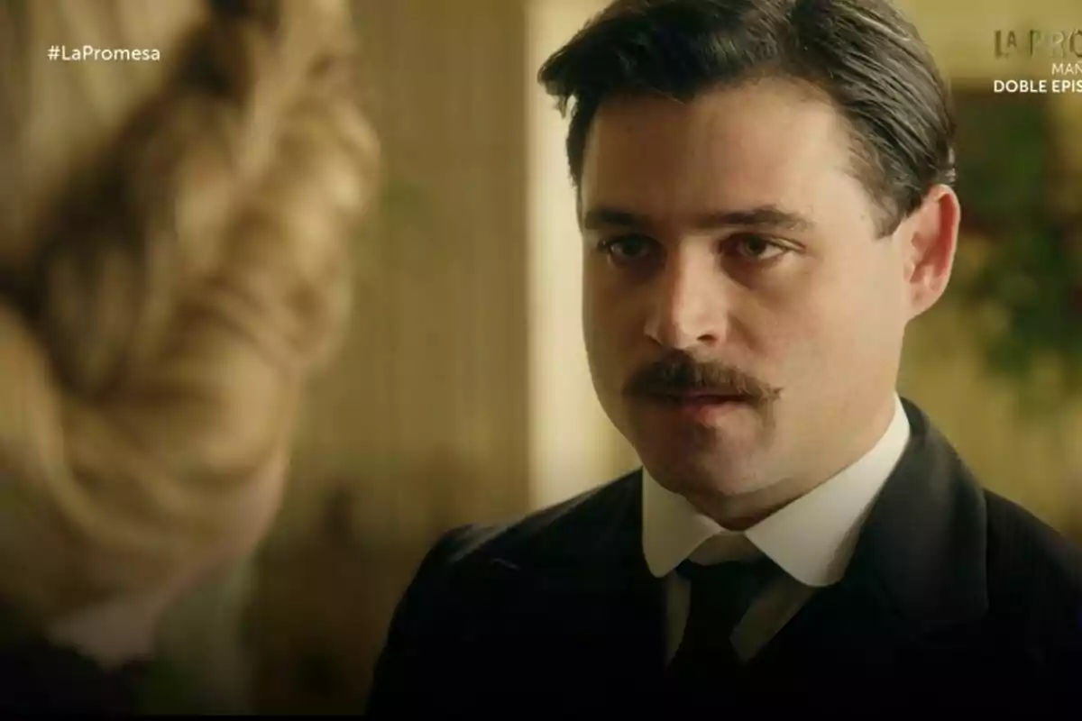 Arturo Sancho con bigote y traje oscuro mira atentamente a una persona de cabello rubio en un entorno interior en La Promesa. Arturo Sancho con bigote y traje oscuro mira atentamente a una persona de cabello rubio en un entorno interior en La Promesa.