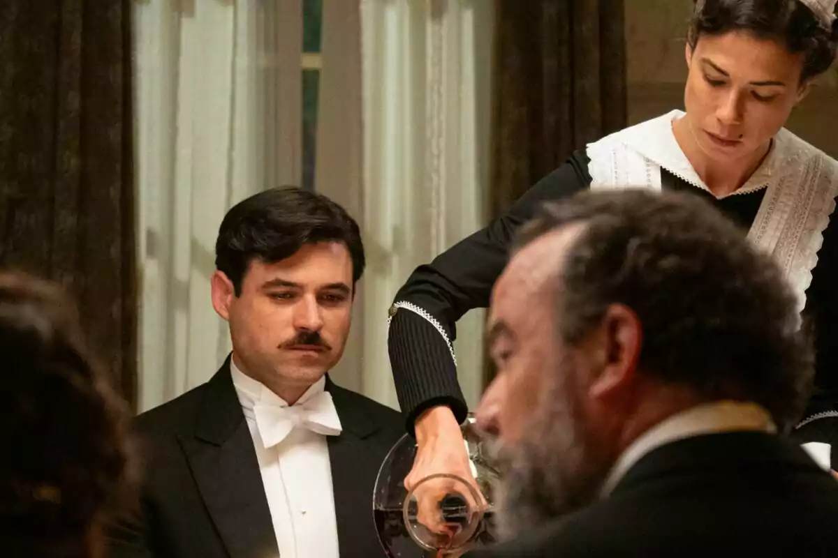 Una camarera sirve vino a Manuel con bigote que está sentado en una mesa elegantemente vestida, mientras otro hombre de barba está en primer plano en La Promesa. Una camarera sirve vino a Manuel con bigote que está sentado en una mesa elegantemente vestida, mientras otro hombre de barba está en primer plano en La Promesa.
