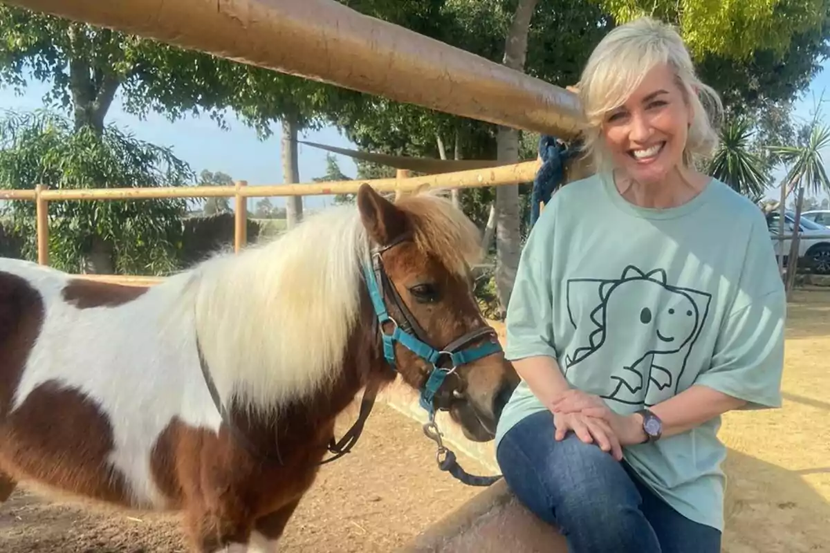 Mar Vega sonriente con una camiseta verde claro con un dibujo de dinosaurio, sentada junto a un pequeño caballo marrón y blanco con una crin rubia, en un entorno al aire libre con árboles y una cerca de madera en Patas Arriba. Mar Vega sonriente con una camiseta verde claro con un dibujo de dinosaurio, sentada junto a un pequeño caballo marrón y blanco con una crin rubia, en un entorno al aire libre con árboles y una cerca de madera en Patas Arriba.