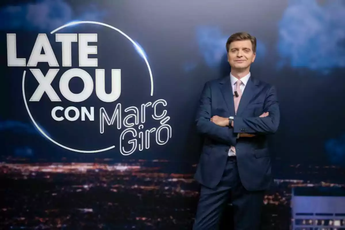 Marc Giró amb un vestit posa amb els braços plegats davant d'un fons que publicita el seu programa, 