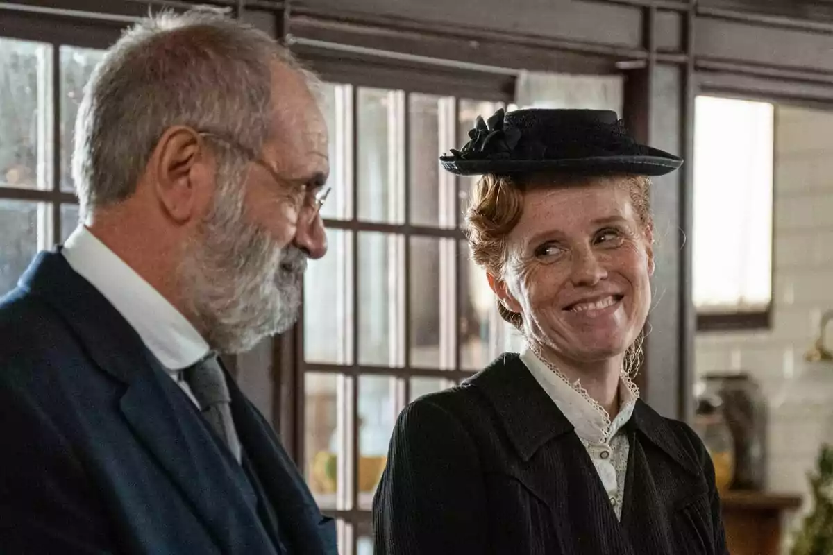Joaquín Climent mayor con barba y María Castro con sombrero sonríen mientras están de pie en una habitación con ventanas grandes en La Promesa. Joaquín Climent mayor con barba y María Castro con sombrero sonríen mientras están de pie en una habitación con ventanas grandes en La Promesa.