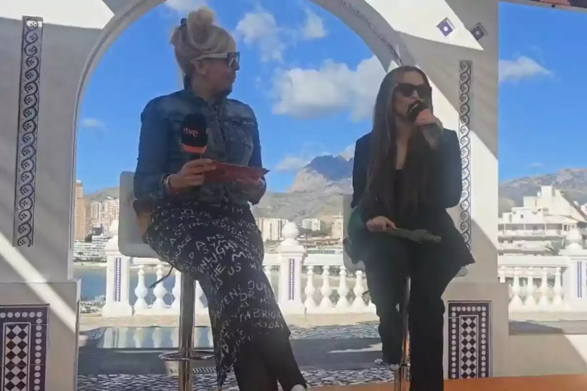 María Eizaguirre y Melody sentadas en un escenario al aire libre con micrófonos en mano y un paisaje urbano y montañoso de fondo en el Benidorm Fest. María Eizaguirre y Melody sentadas en un escenario al aire libre con micrófonos en mano y un paisaje urbano y montañoso de fondo en el Benidorm Fest.