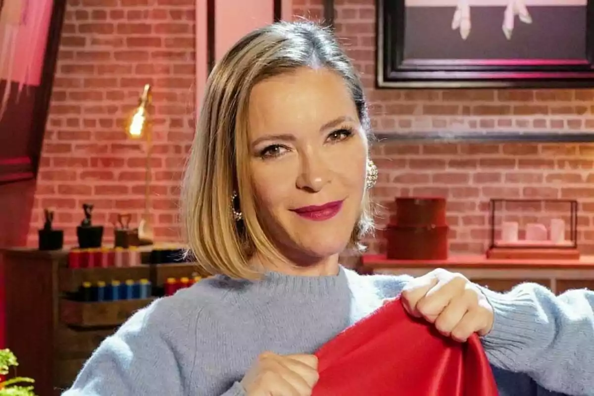 María Esteve con cabello rubio corto sostiene una tela roja en un ambiente interior con paredes de ladrillo y decoración moderna en Maestros de la Costura Celebrity. María Esteve con cabello rubio corto sostiene una tela roja en un ambiente interior con paredes de ladrillo y decoración moderna en Maestros de la Costura Celebrity.