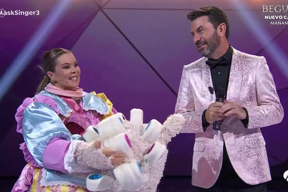 María José Campanario disfrazada con un traje colorido y voluminoso junto a Arturo Valls con un traje brillante en un escenario iluminado en Mask Singer. María José Campanario disfrazada con un traje colorido y voluminoso junto a Arturo Valls con un traje brillante en un escenario iluminado en Mask Singer.