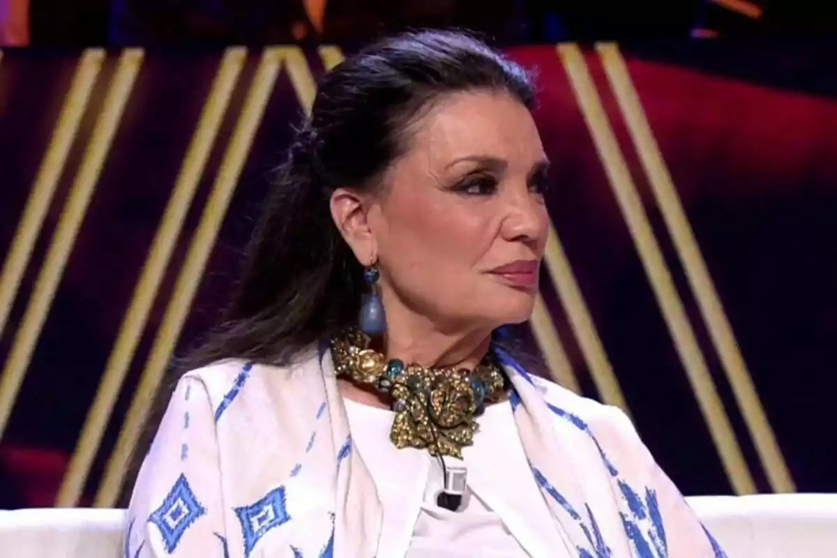 María José Cantudo amb cabell fosc i recollit porta un collaret daurat i blau i una arracada blava, vestida amb una peça blanca amb detalls blaus, asseguda en un entorn il·luminat a De Viernes.