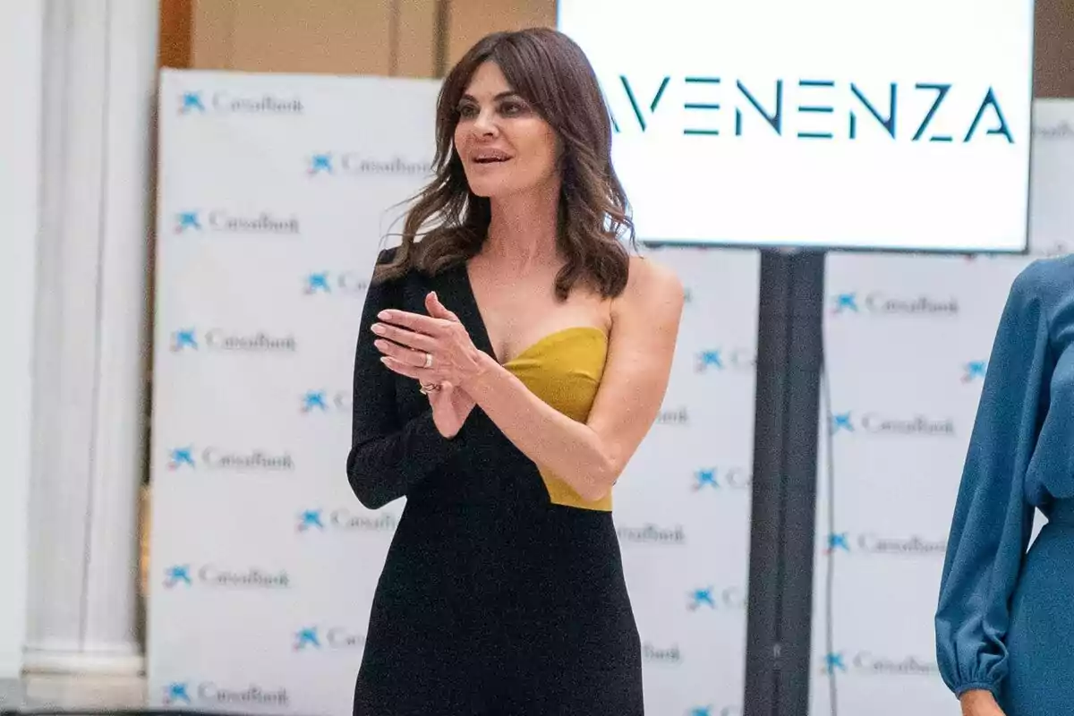 María José Suárez con un vestido negro y amarillo aplaudiendo, con un fondo que muestra logotipos de CaixaBank y un cartel que dice María José Suárez con un vestido negro y amarillo aplaudiendo, con un fondo que muestra logotipos de CaixaBank y un cartel que dice