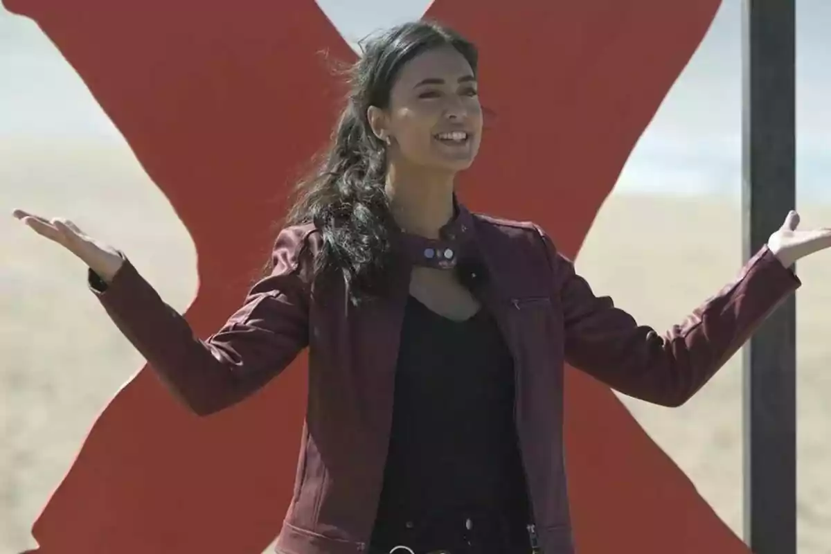 Maria Lamela somrient amb jaqueta vermella i els braços estesos davant d'una gran X vermella a la platja. Maria Lamela somrient amb jaqueta vermella i els braços estesos davant d'una gran X vermella a la platja.