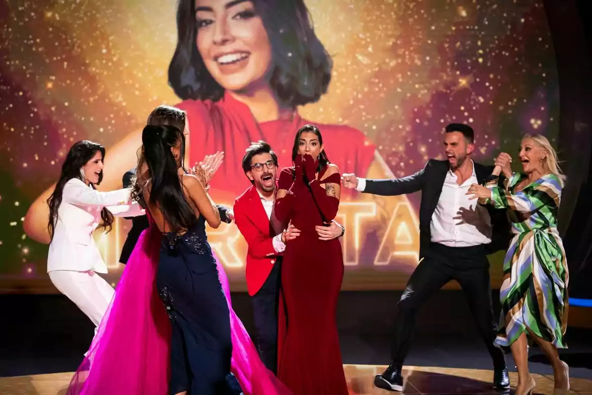 Un grup de persones format per Marieta Díaz, Dani Santos, Suso Álvarez, Jeimy Báez, Maica Benedicto o María Sánchez, celebra emocionat en un escenari amb una imatge de fons d'una dona somrient a GH DÚO.