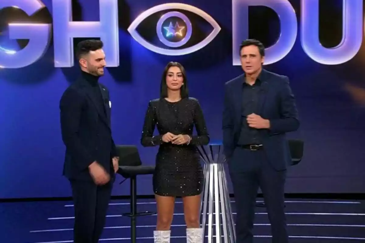 Suso Álvarez, Marieta Díaz y Ion Aramendi de pie en un set de televisión con un fondo que tiene un logotipo de ojo y letras grandes de GH DÚO. Suso Álvarez, Marieta Díaz y Ion Aramendi de pie en un set de televisión con un fondo que tiene un logotipo de ojo y letras grandes de GH DÚO.