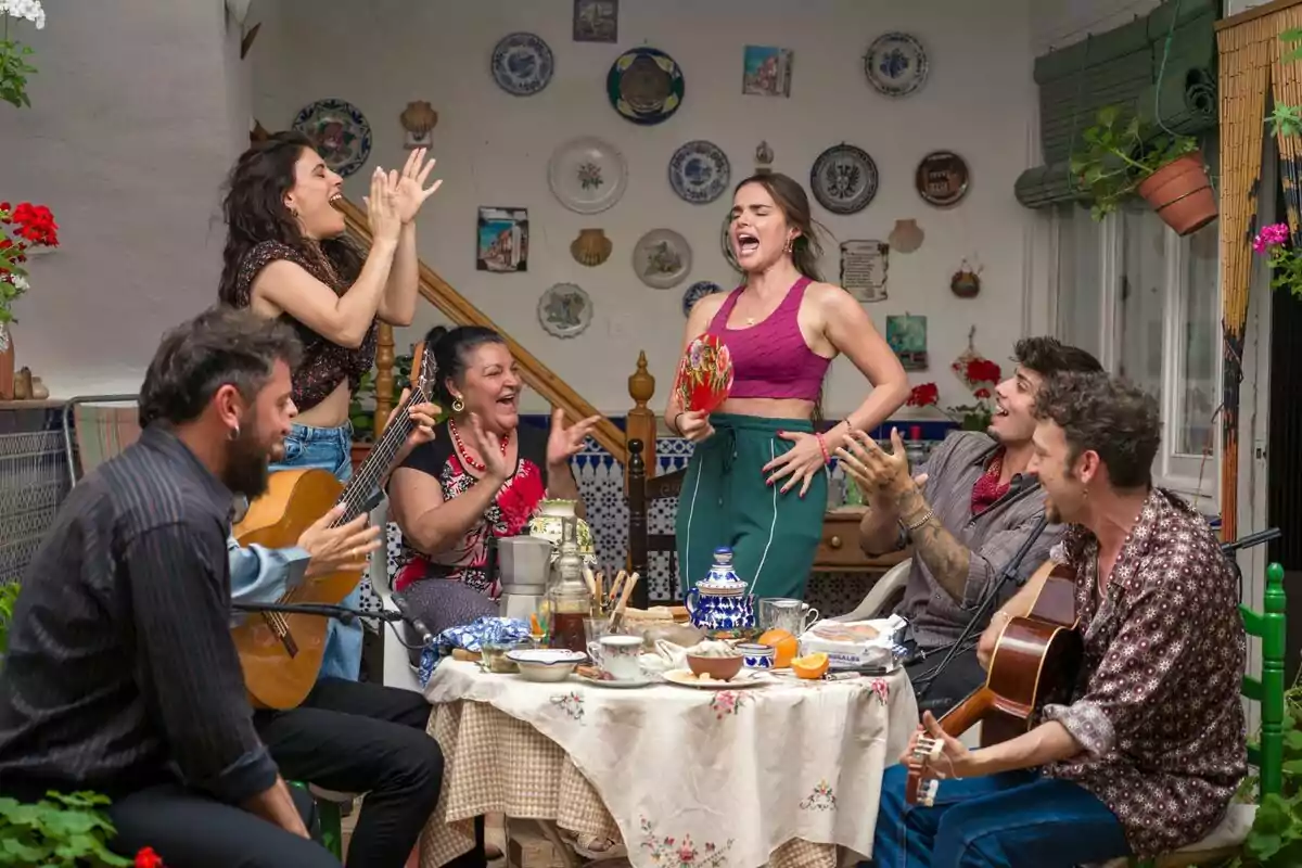 Un grupo de personas con Melody como protagonista disfruta de una reunión animada en un patio decorado con platos en la pared, mientras tocan la guitarra, cantan y aplauden alrededor de una mesa con comida y bebida en Mariliendre.