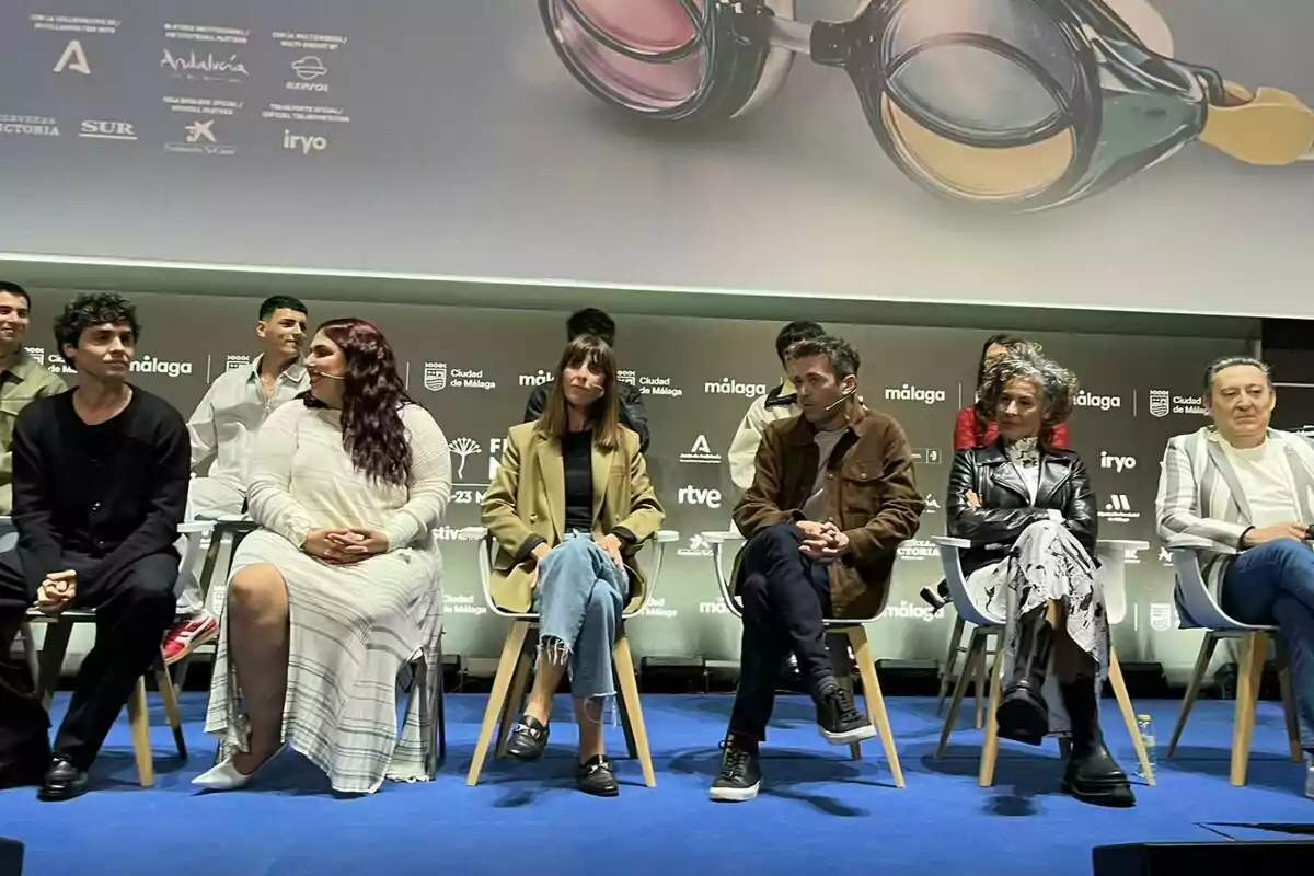 Un grupo de personas sentadas en sillas sobre un escenario durante un evento, con un fondo que muestra logotipos y una imagen de gafas en Mariliendre.