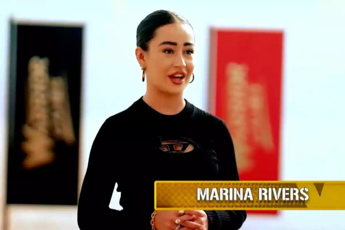Marina Rivers de cabello oscuro recogido y camiseta negra hablando frente a dos cuadros desenfocados con un recuadro amarillo que muestra el nombre Marina Rivers en 'Warrior Games'.