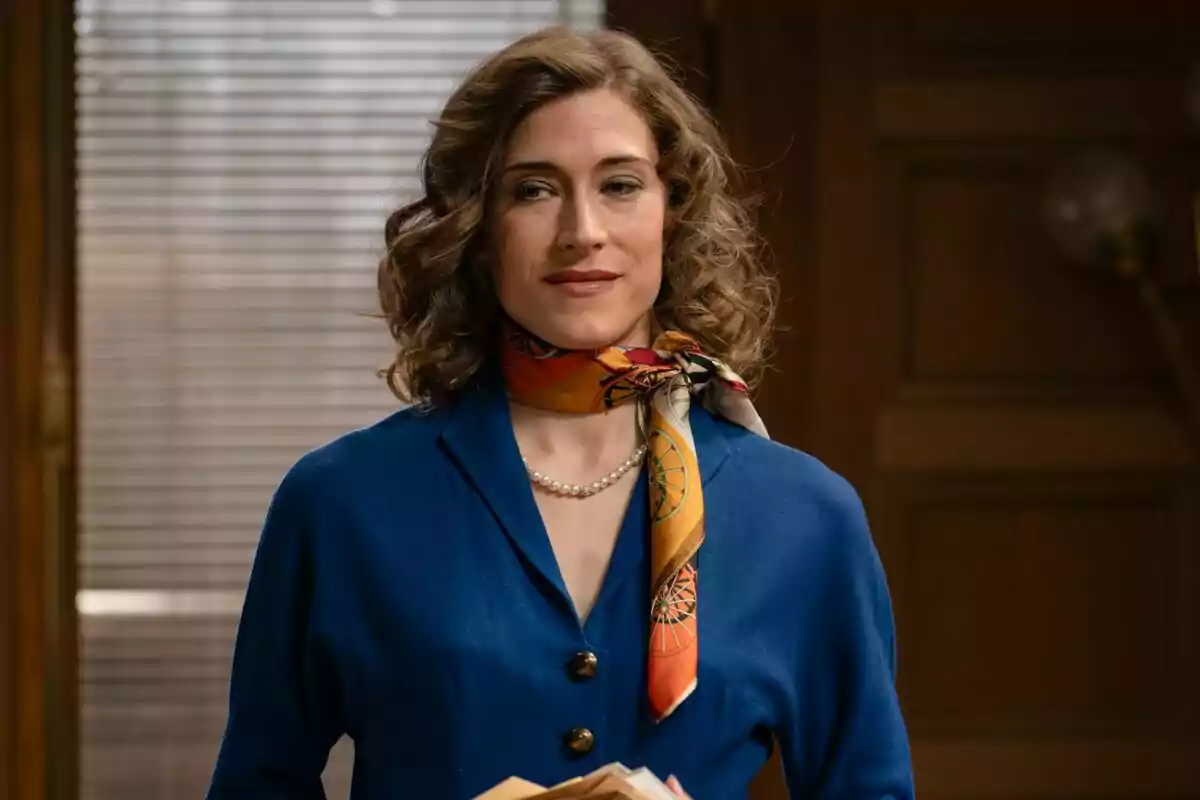 Captura de Marta Belmonte amb cabell arrissat i castanyer, vestida amb un vestit blau i un mocador colorit al voltant del coll, dret en una habitació amb una finestra de persianes al fons en Sueños de Libertad Captura de Marta Belmonte amb cabell arrissat i castanyer, vestida amb un vestit blau i un mocador colorit al voltant del coll, dret en una habitació amb una finestra de persianes al fons en Sueños de Libertad