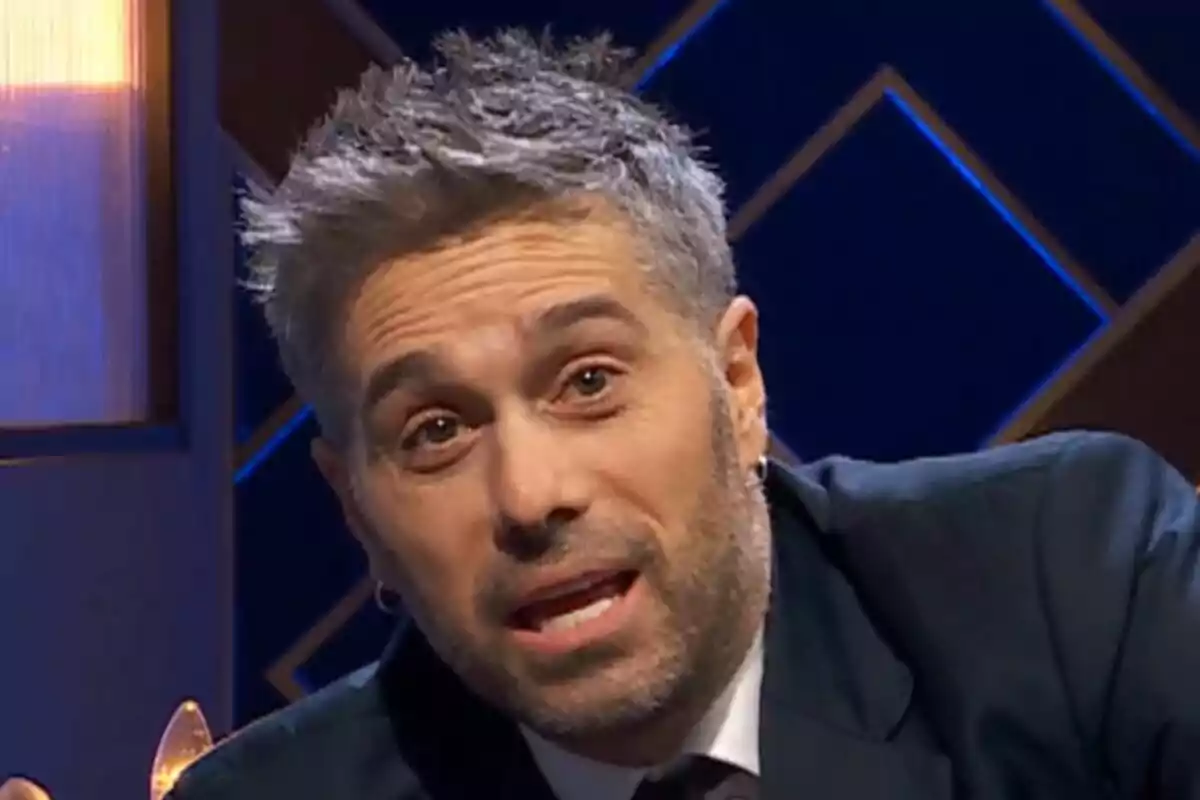 Dani Martínez con cabello corto y barba está hablando en un entorno iluminado con luces azules y doradas en Martínez y Hermanos.