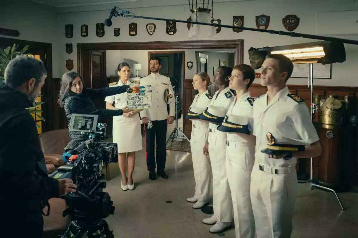 Un grupo de personas encabezado por Lucía Jiménez y Alfonso Bassave vestidas con uniformes navales está en un set de filmación, con cámaras y equipo de grabación alrededor, mientras una persona sostiene una claqueta en la serie Marusía: Vientos de Honor. Un grupo de personas encabezado por Lucía Jiménez y Alfonso Bassave vestidas con uniformes navales está en un set de filmación, con cámaras y equipo de grabación alrededor, mientras una persona sostiene una claqueta en la serie Marusía: Vientos de Honor.