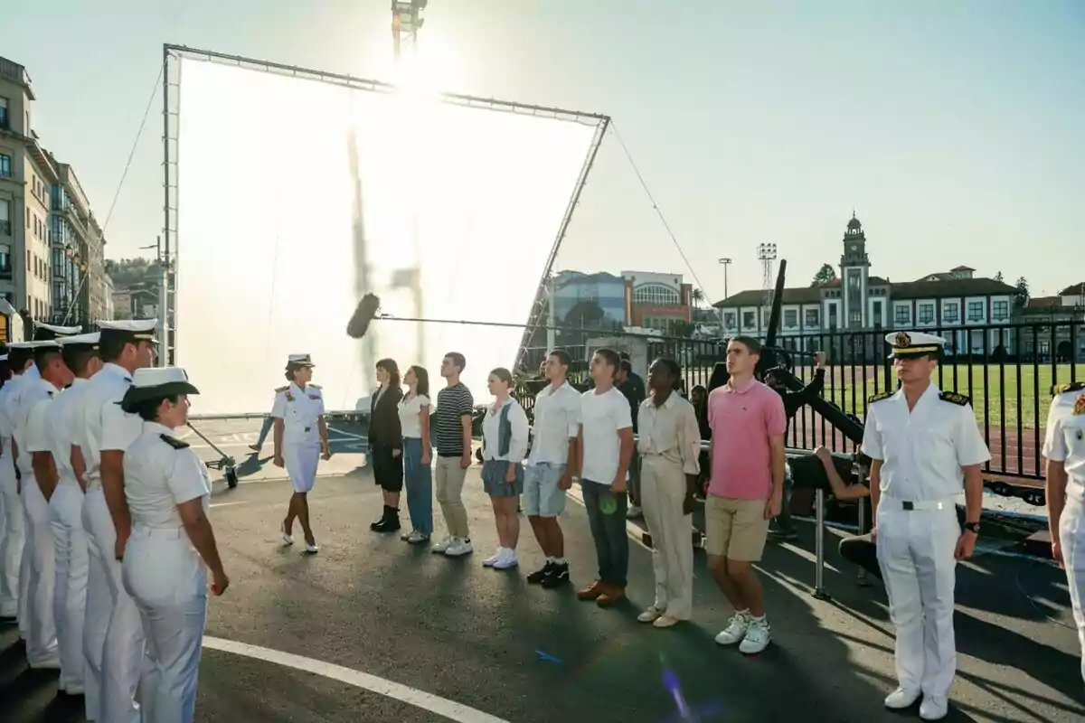 Un grupo de personas vestidas con uniformes navales y ropa casual están alineadas en una calle al aire libre, con un gran reflector detrás de ellas y un micrófono de boom visible, sugiriendo que se está filmando una escena en la serie Marusía: Vientos de Honor. Un grupo de personas vestidas con uniformes navales y ropa casual están alineadas en una calle al aire libre, con un gran reflector detrás de ellas y un micrófono de boom visible, sugiriendo que se está filmando una escena en la serie Marusía: Vientos de Honor.