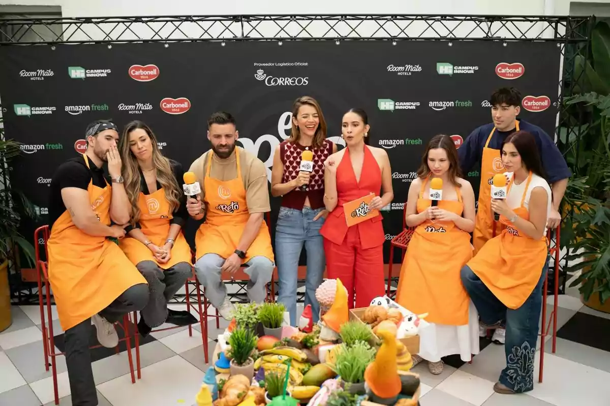 Un grupo de personas con delantales naranjas y micrófonos se encuentra frente a un fondo con logotipos de marcas, mientras una mesa con frutas y decoraciones está en primer plano en la presentación de 'El Reto' de 'Masterchef''. Un grupo de personas con delantales naranjas y micrófonos se encuentra frente a un fondo con logotipos de marcas, mientras una mesa con frutas y decoraciones está en primer plano en la presentación de 'El Reto' de 'Masterchef''.