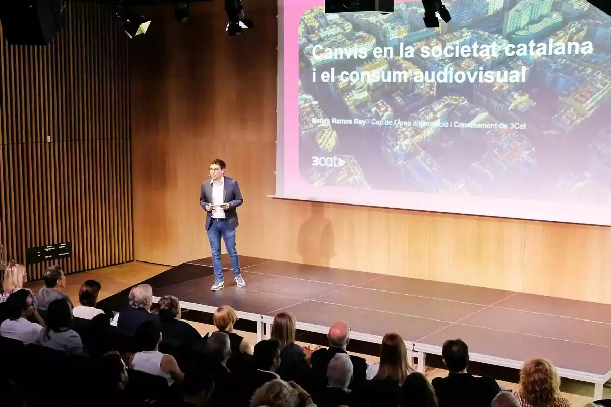 Maties Ramos dret damunt d’un escenari fa una presentació davant d’una audiència, amb una pantalla gran al fons que mostra un títol sobre els canvis en la societat catalana i el consum audiovisual de 3Cat.