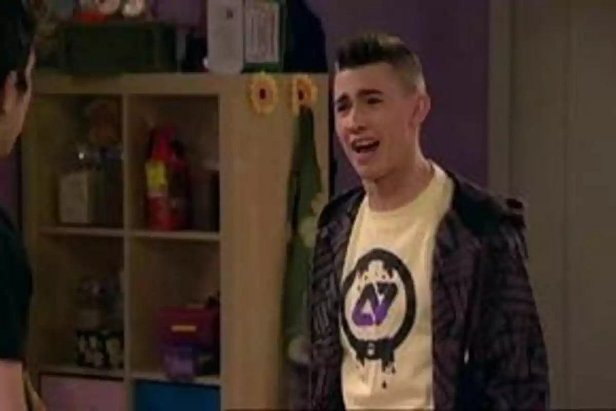 Adrián Gordillo con una camiseta clara y chaqueta oscura está hablando en una habitación con estanterías al fondo en Aída. Adrián Gordillo con una camiseta clara y chaqueta oscura está hablando en una habitación con estanterías al fondo en Aída.