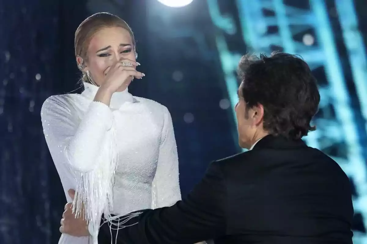 Melani emocionada con un vestido blanco es consolada por Manel Fuentes en un escenario iluminado de Tu Cara Me Suena. Melani emocionada con un vestido blanco es consolada por Manel Fuentes en un escenario iluminado de Tu Cara Me Suena.