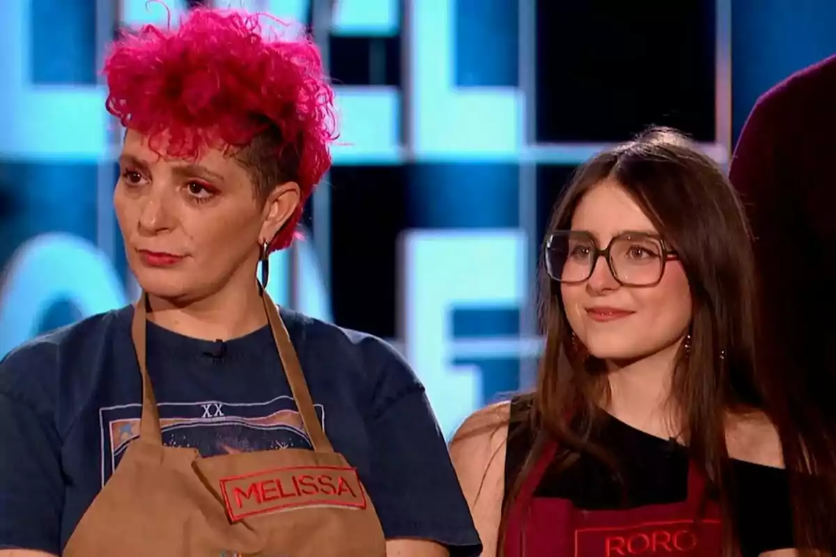 Dues persones dretes, la Melissa amb cabell arrissat i tenyit de rosa i en Roro Bueno amb cabell llarg i ulleres, ambdues porten davantals amb noms brodats a Next Level Chef.