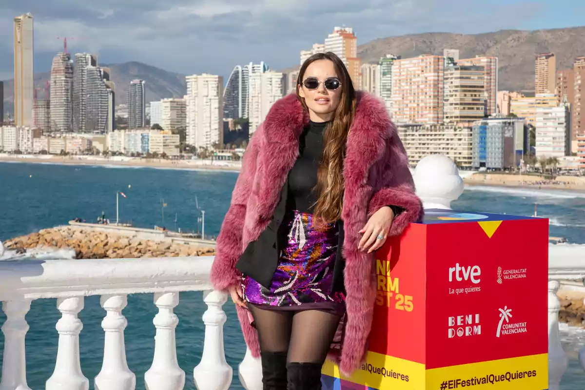 Melody con abrigo de piel rosa y gafas de sol posa frente a un paisaje urbano costero con edificios altos y el mar de fondo. Melody con abrigo de piel rosa y gafas de sol posa frente a un paisaje urbano costero con edificios altos y el mar de fondo.