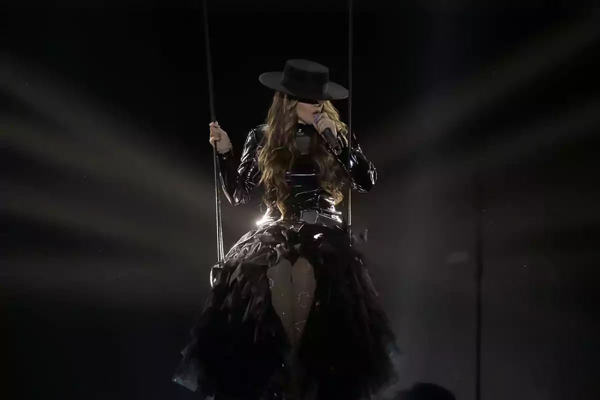 Melody con sombrero negro y vestido oscuro canta mientras está sentada en un columpio iluminado por un foco en el escenario del Benidorm Fest. Melody con sombrero negro y vestido oscuro canta mientras está sentada en un columpio iluminado por un foco en el escenario del Benidorm Fest.