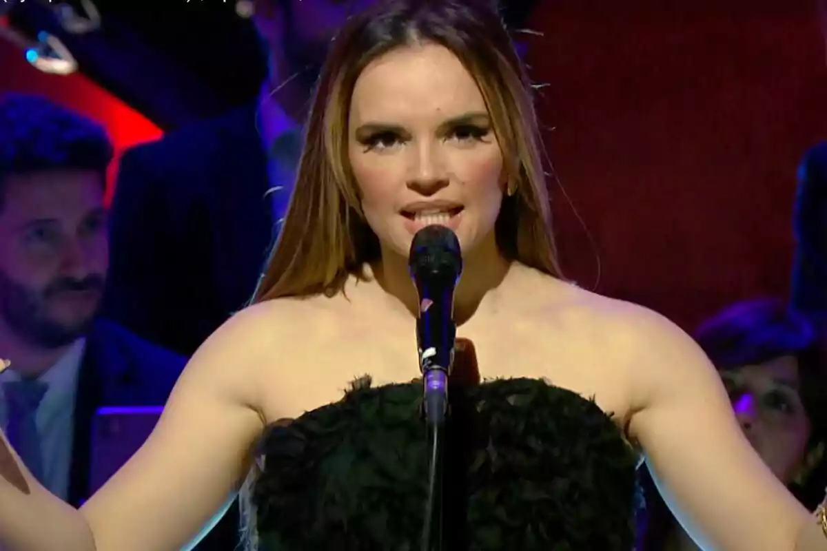 Melody canta en un escenario con un micrófono acompañada de la Orquesta Sinfónica de RTVE, vestida de negro, mientras el fondo está iluminado con luces de colores.