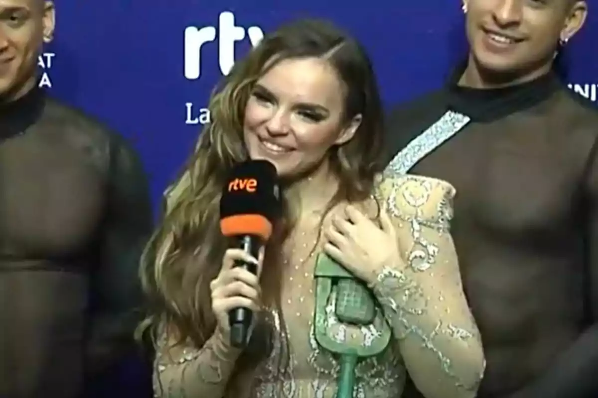 Melody sonriente sostiene un micrófono mientras está acompañada por dos personas en un evento con fondo azul en la rueda de prensa tras el Benidorm Fest.