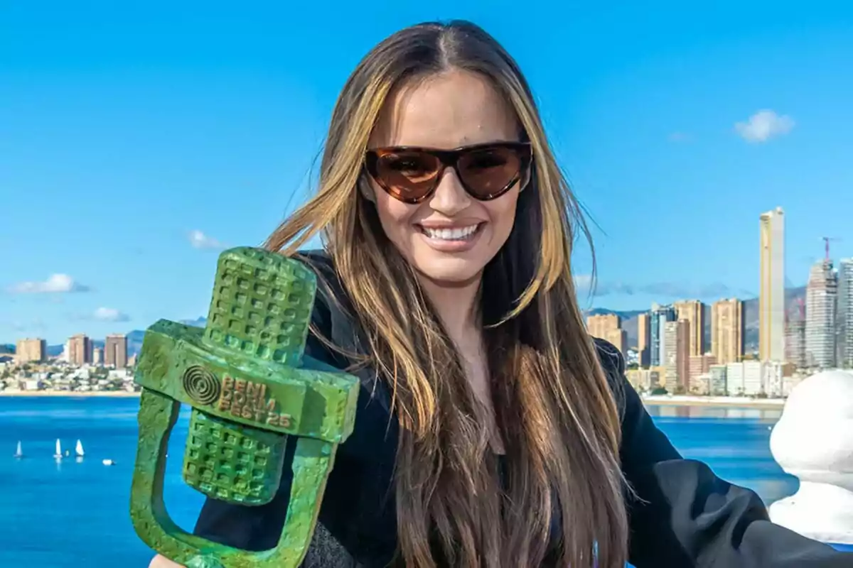 Melody con gafas de sol sonríe mientras sostiene un trofeo verde con la inscripción "Benidorm Fest 23" frente a un paisaje urbano y marino.