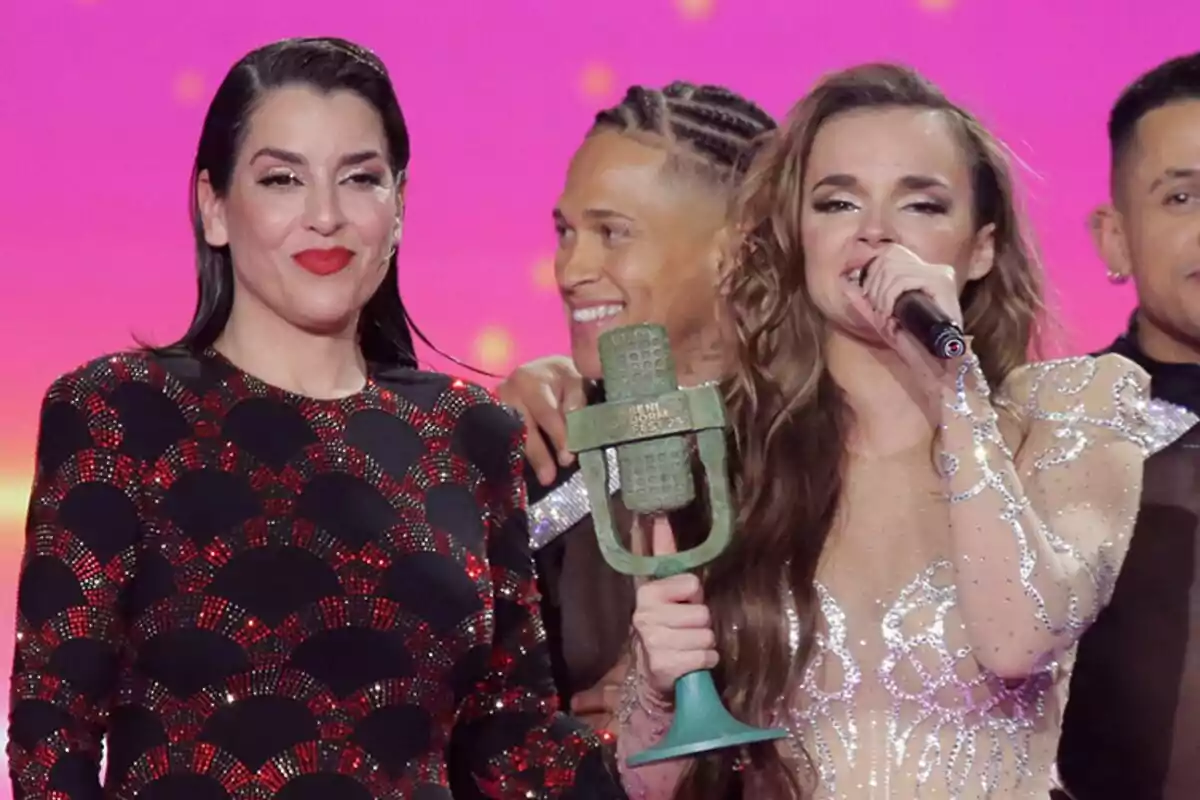 Un grup de persones en un escenari del Benidorm Fest, Melody sosté un trofeu i un micròfon al costat de Ruth Lorenzo, amb un fons de color rosa brillant.
