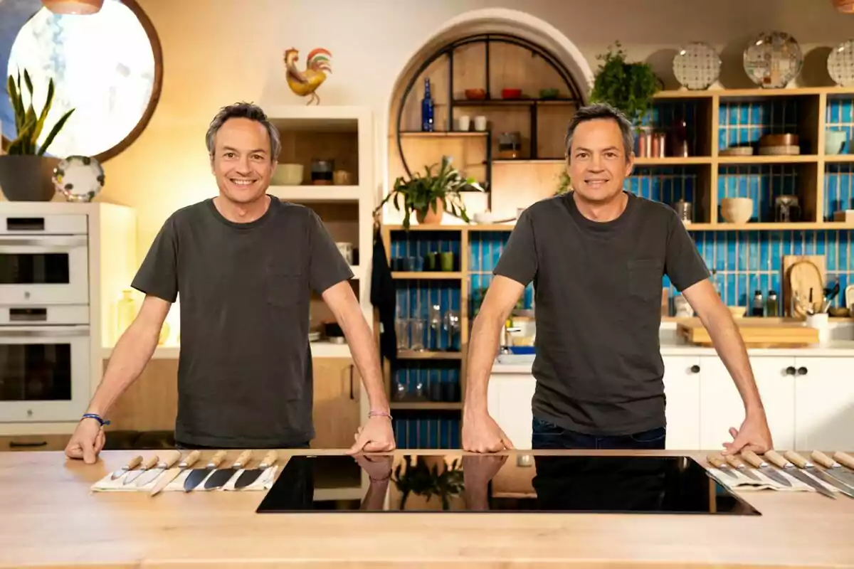 Los Hermanos Torres de pie en una cocina moderna con encimera de madera y cuchillos alineados, rodeados de estantes con utensilios y decoración. Los Hermanos Torres de pie en una cocina moderna con encimera de madera y cuchillos alineados, rodeados de estantes con utensilios y decoración.