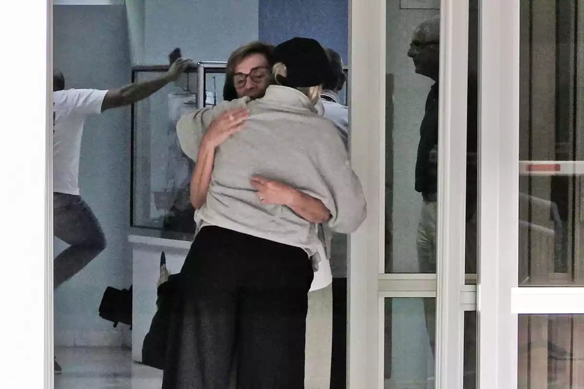 Mercedes Bernal i una altra noia s'abracen afectuosament en un espai interior mentre altres persones es veuen reflectides al vidre.