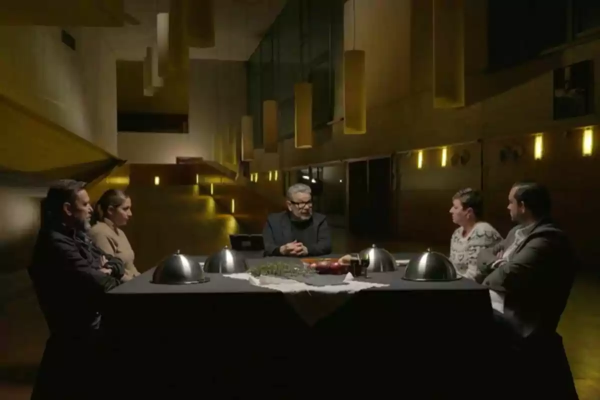 Cinco personas sentadas alrededor de una mesa en una habitación con iluminación tenue y lámparas colgantes en 'Batalla de restaurantes'. Cinco personas sentadas alrededor de una mesa en una habitación con iluminación tenue y lámparas colgantes en 'Batalla de restaurantes'.